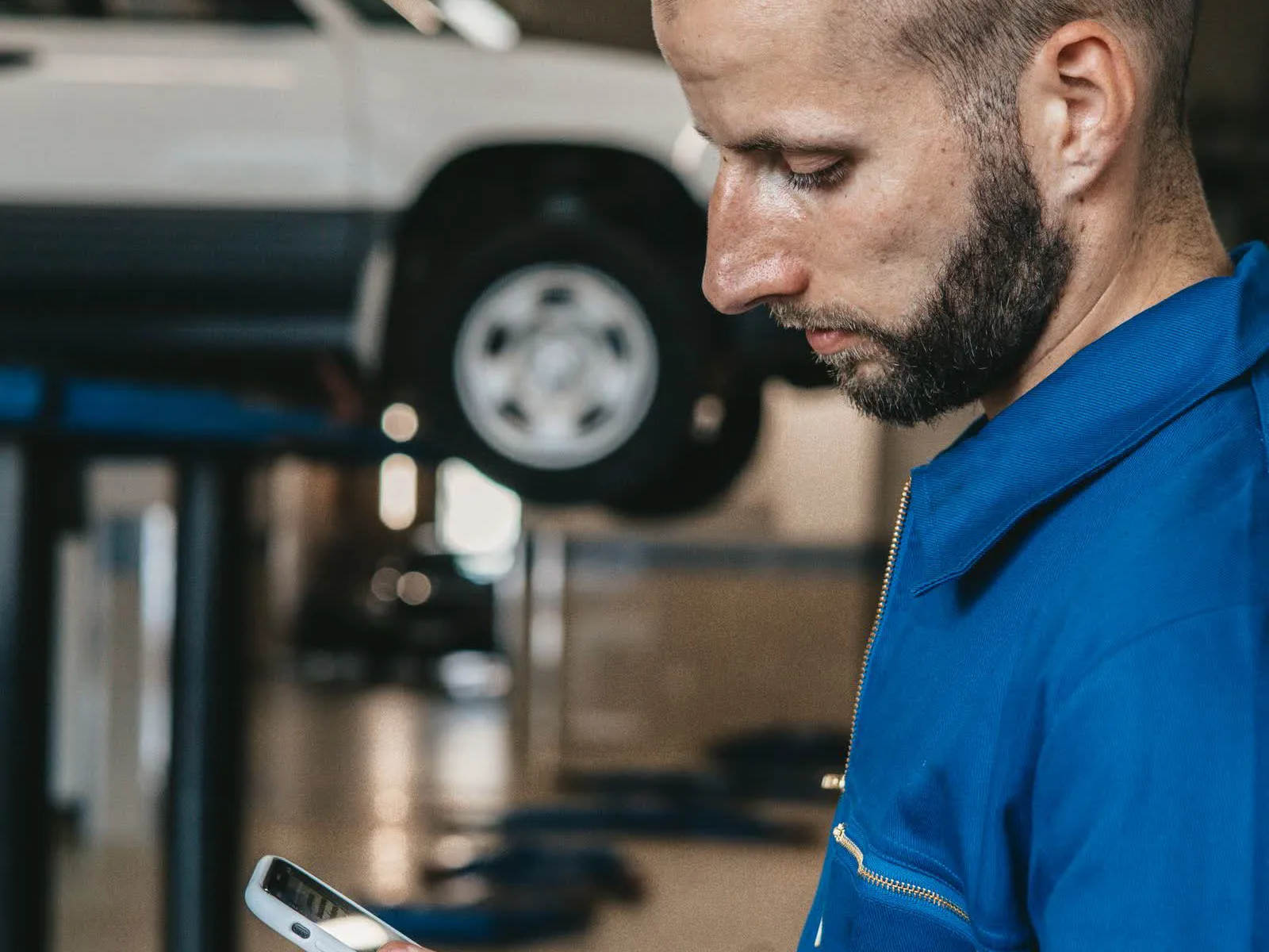Mechanik w niebieskim kombinezonie sprawdza coś w telefonie, za nim samochód na podnośniku w warsztacie