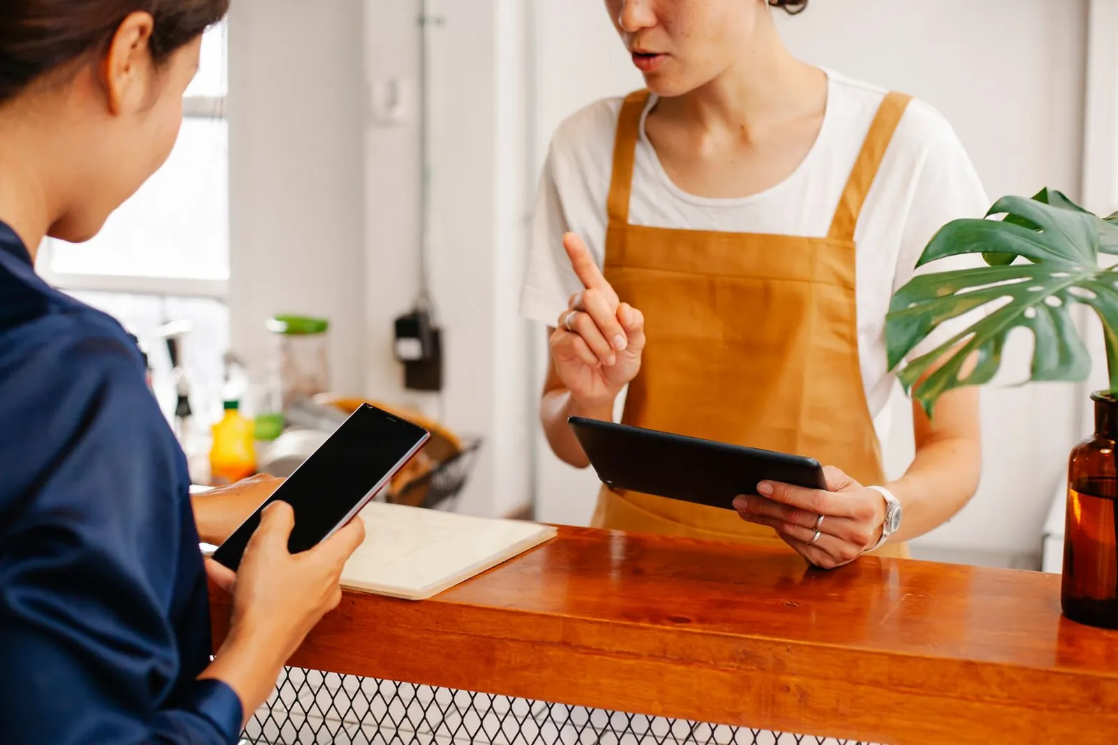 A customer holds her phone at a shop counter while the owner in an apron looks at a tablet