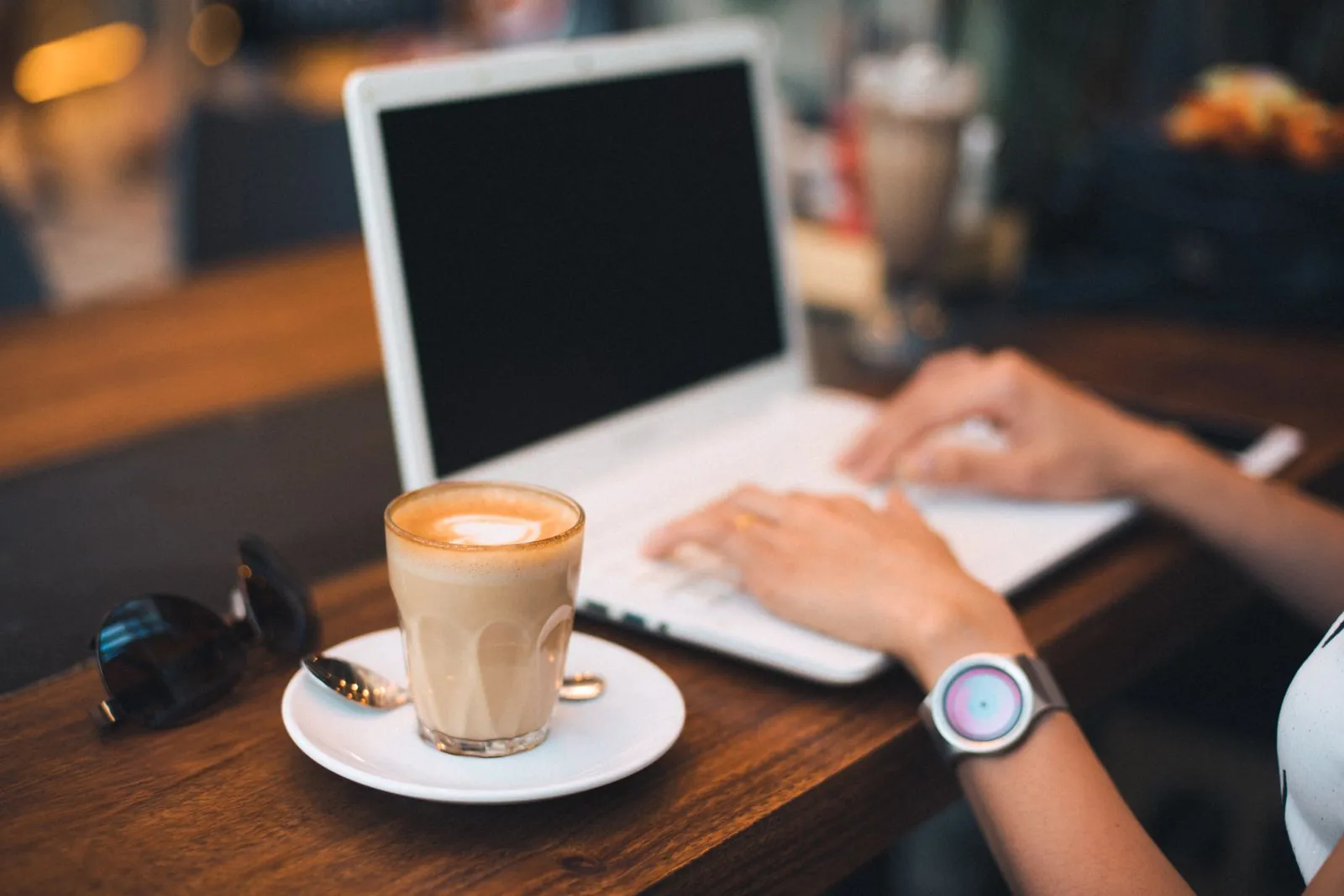 Hands typing on a laptop next to a latte on a wooden café table