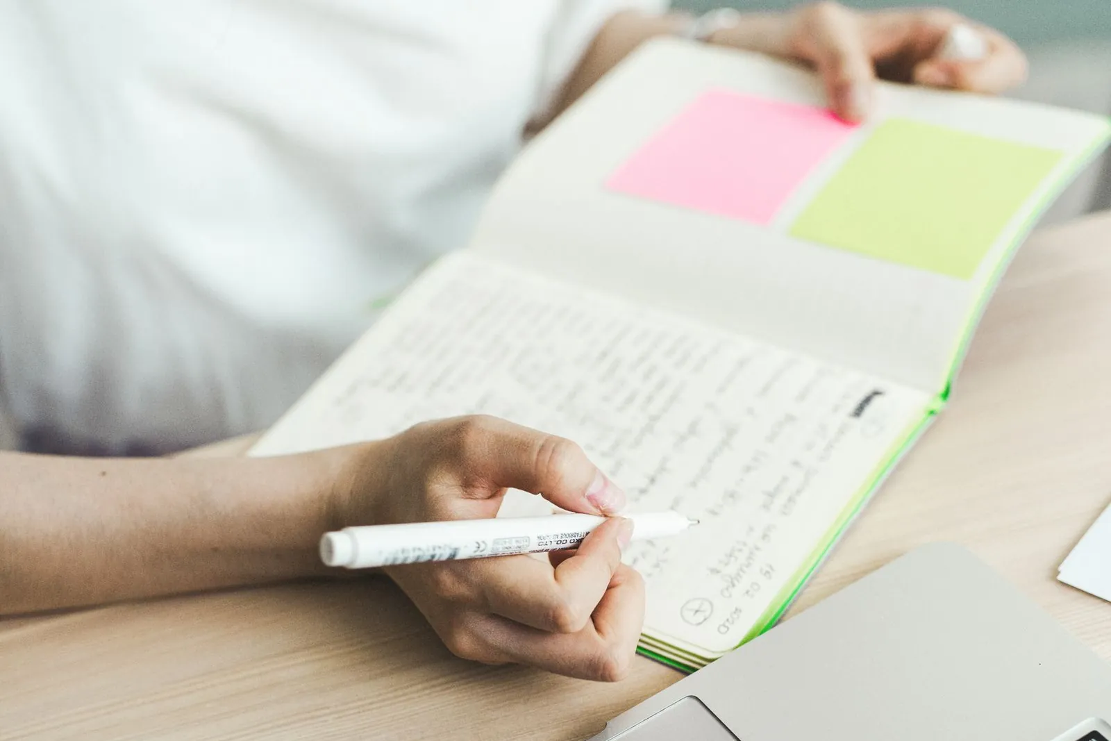 A hand writes in a notebook with sticky notes, a laptop beside it