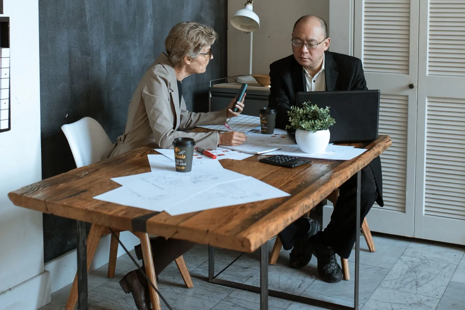Two people at a wooden table with papers, a laptop and a phone go over a plan