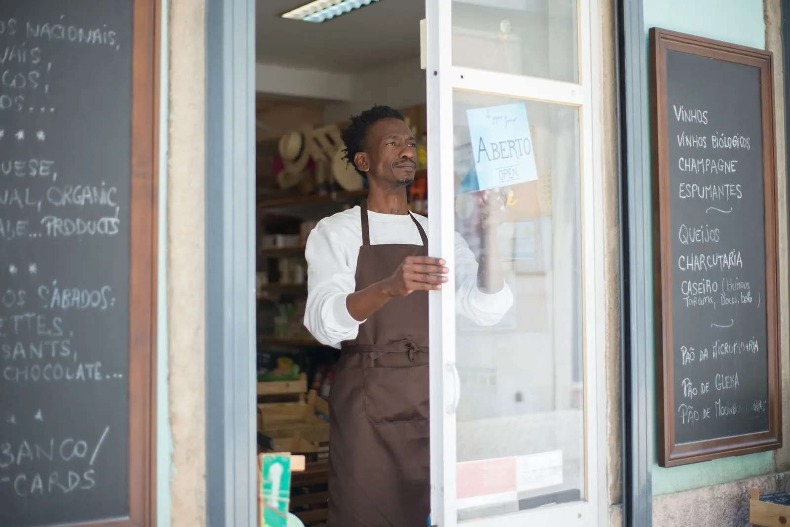 A shop owner in an apron holds open the door of his small grocery store