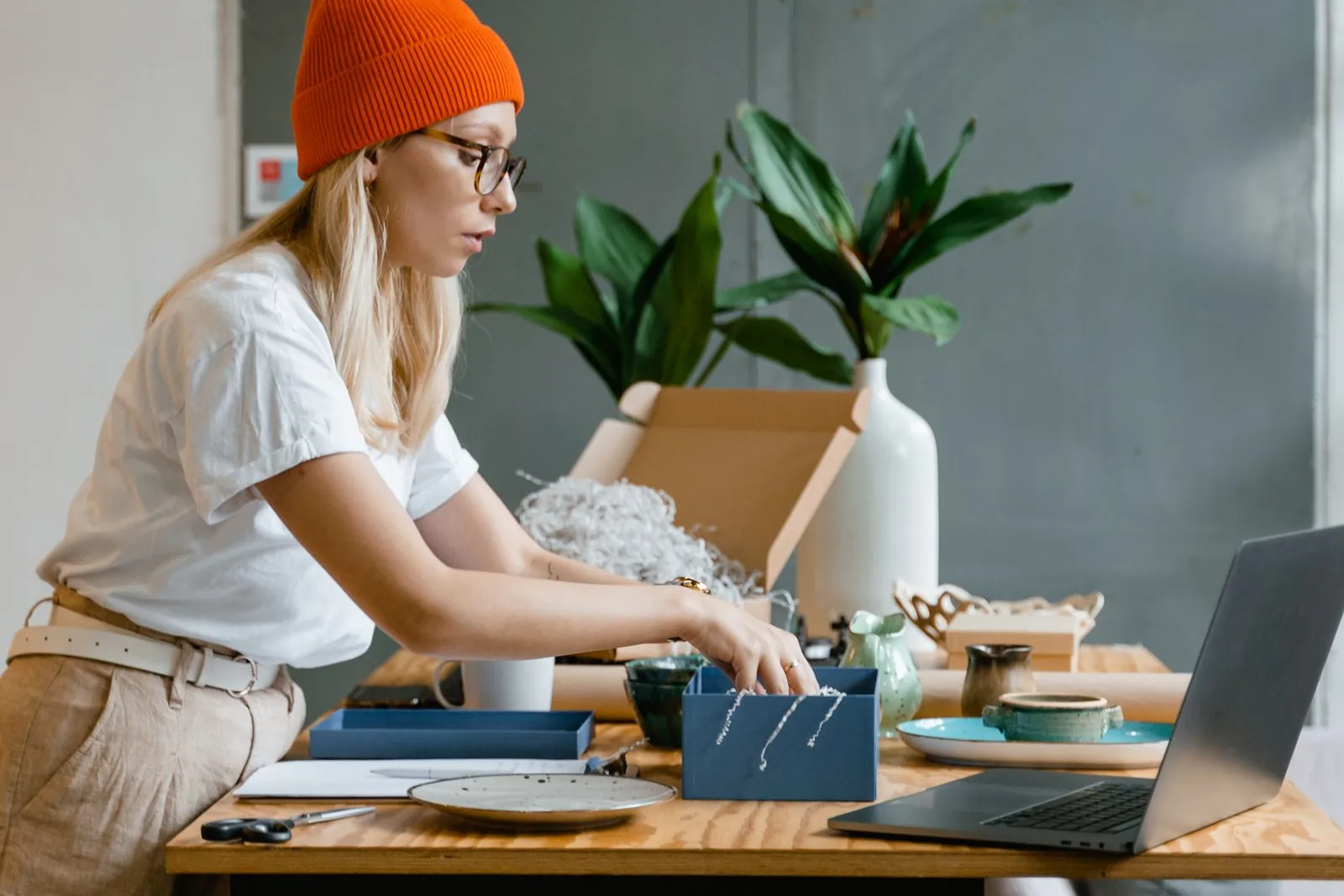 A woman in an orange beanie packs a handmade order at a workbench with a laptop