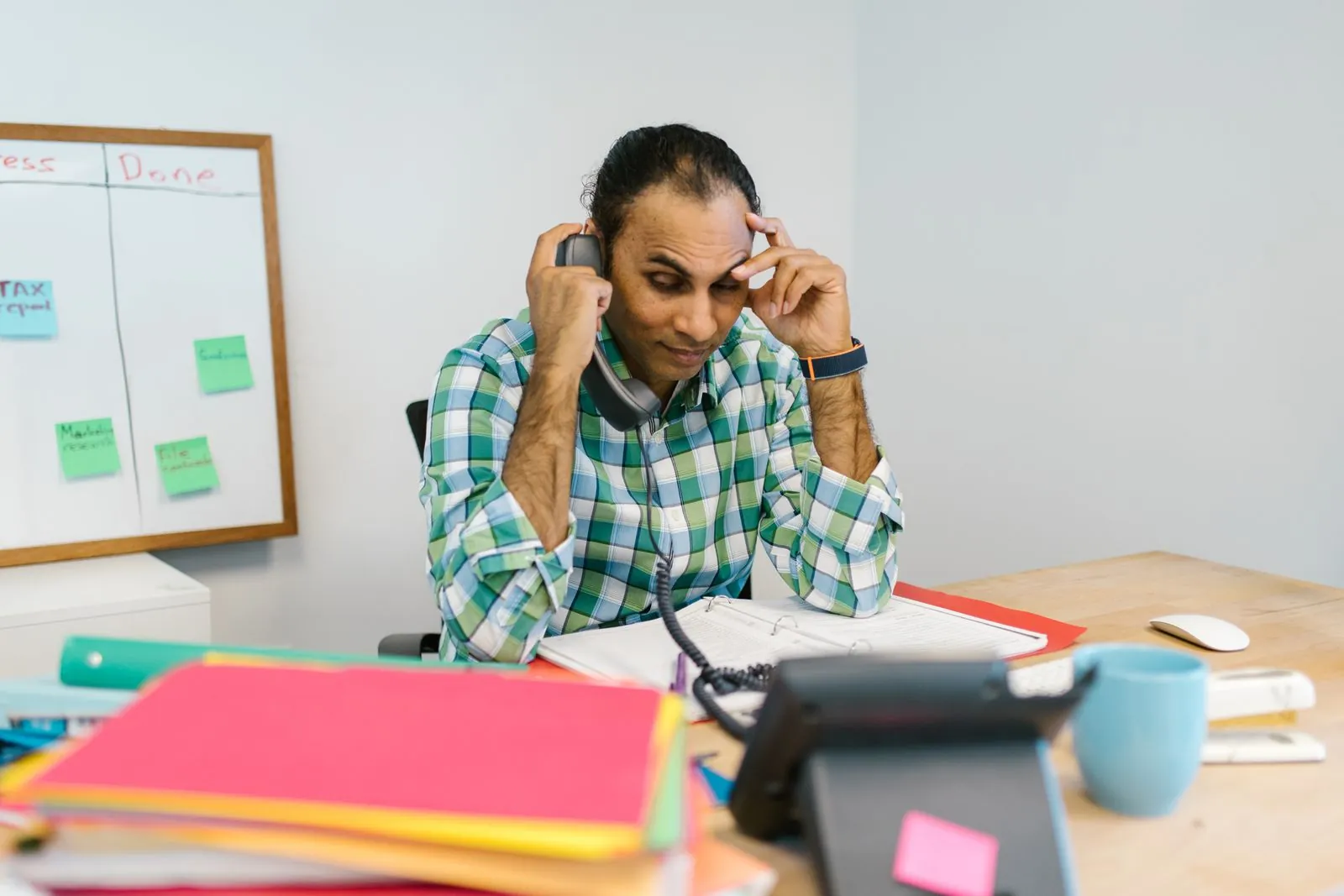 A man at a cluttered desk rests his head on his hand over paperwork, phone at his ear